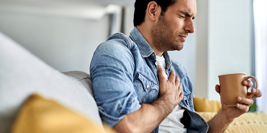 Young man feeling sick and holding his chest in pain while drinking tea in the living room.