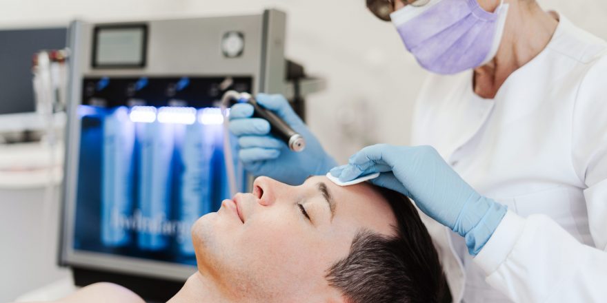 Senior, professional doctor, cosmetologist, woman doing hydra facial, mesotherapy, microdermabrasion peeling to young handsome man, in modern cosmetic clinic. Concept of cosmetology, rejuvenation